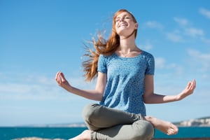 Beautiful woman meditating at the seaside sitting cross-legged on the rocks with her head tilted to the sun and her eyes closed in ecstasy