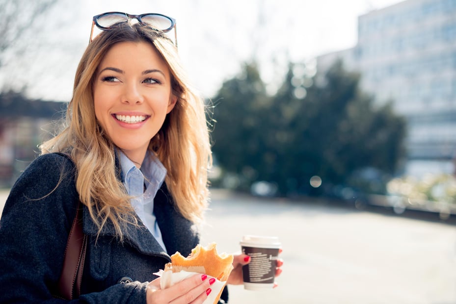 A female employee walks across the street while holding a breakfast sandwich and coffee.