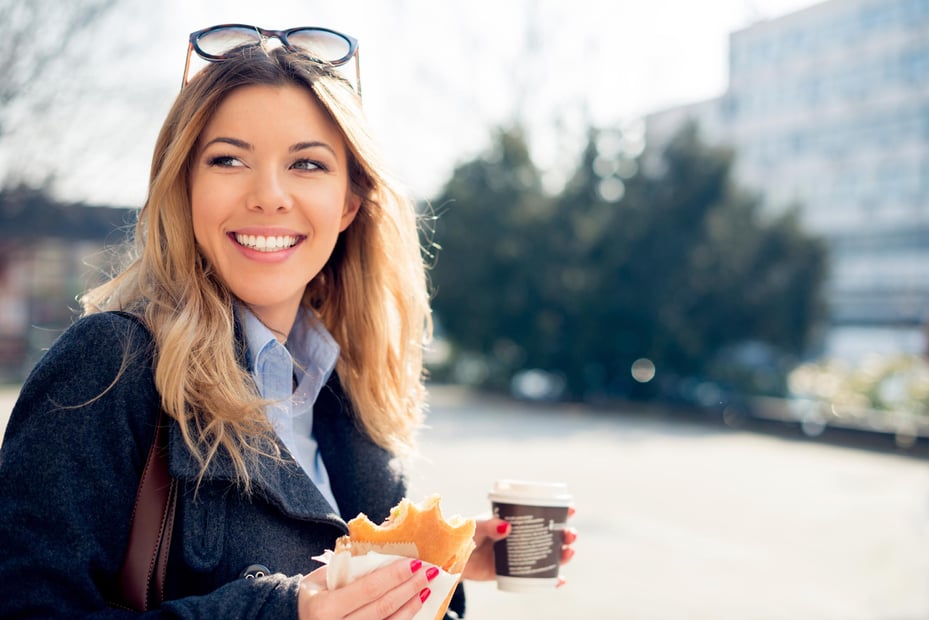 A female employee walks across the street while holding a breakfast sandwich and coffee. 