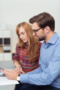 Business man and woman working on a tablet computer as they sit side by side at a desk in the office, closeup side view