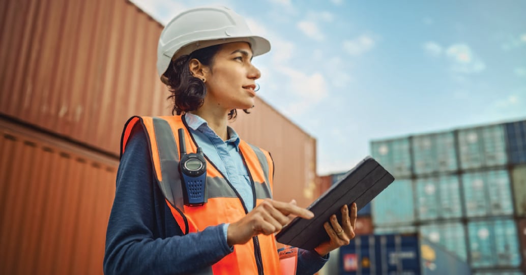 A female employee wearing PPE uses a tablet while standing on a loading dock.